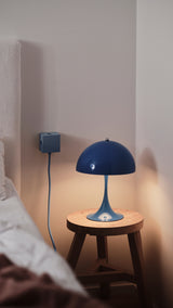 AVOLT Square 1 USB-C charger (CH) in blue mounted beside a bed, cable to a lit blue table lamp on a wooden stool.