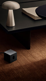 AVOLT Square 1 USB-C Swiss cube charger, black, on brown carpet beneath table beside lamp and stacked papers.