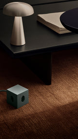 AVOLT Square 1 (CH) USB-C green cube outlet on carpet beside black low table, lamp and stacked paper
