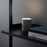 lifestyle: AVOLT Lamp 1 Gotland gray cube lamp glowing on a shelf next to books, warm top light