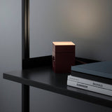 Mulberry red AVOLT Lamp 1 on black desk, cube lamp lit with side outlet beside stacked books