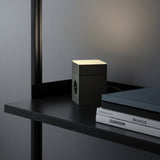 AVOLT Lamp 1 table lamp in oak green glowing on a black shelf beside books and a closed laptop