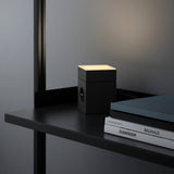 AVOLT Lamp 1 in black on a shelf, warm top light beside books and laptop in a minimalist workspace