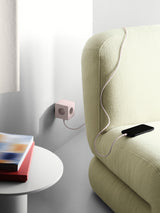 AVOLT Square 2 USB-C charging cube in soft pink plugged into wall, cable to phone on pale green sofa