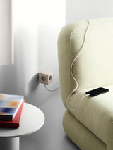 AVOLT Square 2 USB‑C sienas kubs smilškrāsā uzlādē telefonu blakus gaiši zaļai dīvānai.